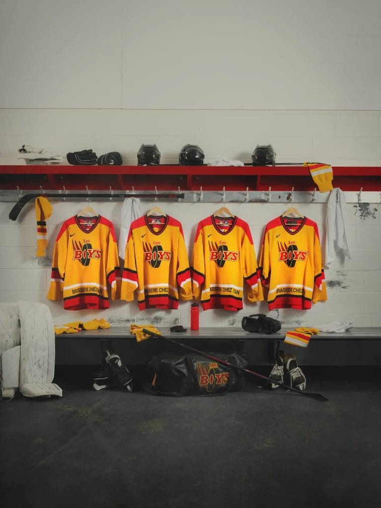 Quatre chandails de hockey jaunes et rouges sont suspendus dans le vestiaire des Boys. En dessous, des casques, des gants et des bâtons sont éparpillés sur un banc et au sol. Un sac de hockey noir est ouvert à l'avant.